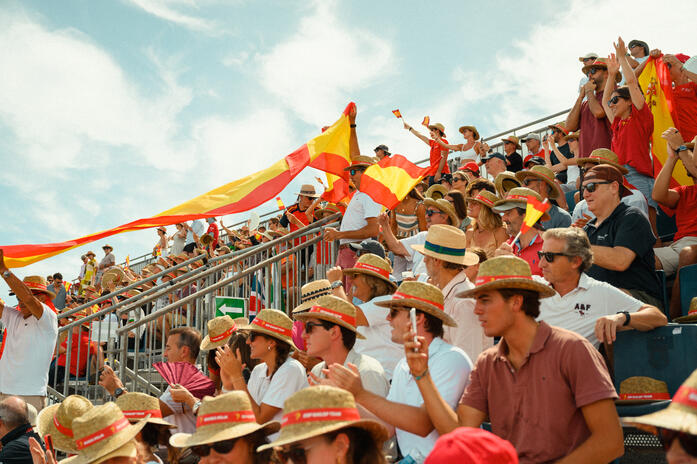 The crowd brings its own energy to the Spain Sail Grand Prix. Spectators cheering and waving Spanish flags in packed grandstands at SailGP in Valencia.