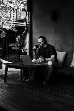 Recording in his working environment Man wearing headphones and speaking into a microphone while working on a laptop outdoors