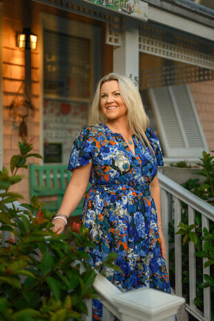 Confidence in colour Smiling blonde woman in a blue floral dress standing outdoors beside a porch