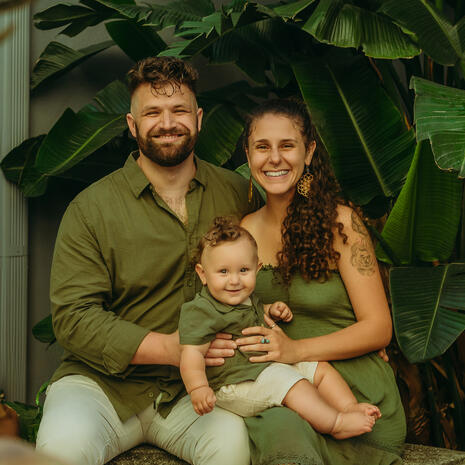 A family portrait in shades of green, full of smiles. Two smiling parents hold their young child on their laps, wearing coordinated green outfits against large tropical leaves.