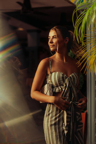 Warm sunlight, palm leaves and a trace of rainbow. A woman in a striped outfit looks to the side beside palm leaves, with warm sunlight on her face and rainbow flare across the foreground.