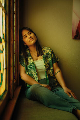 A quiet moment beside a stained-glass window. A woman in a green floral shirt and jeans sits beside a stained-glass window, resting her head against its wooden frame.
