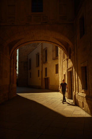 A portrait framed by stone, sunlight and deep shadows. A man in dark clothing and sunglasses walks beneath a large stone arch, with warm sunlight casting long shadows across the pavement.