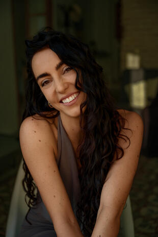 An open smile in soft light. Close portrait of a smiling woman with long dark curls, leaning forward and looking at the camera against a muted green background.