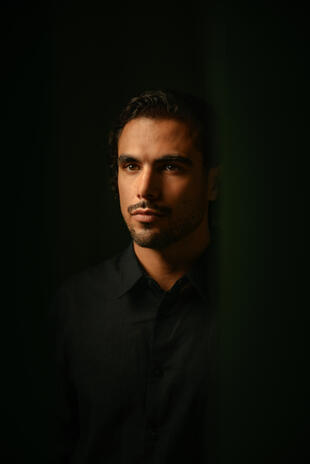 A quiet portrait shaped by warm light and deep green shadows. A man in a black shirt looks slightly away from the camera, his face lit from one side against a dark green background.