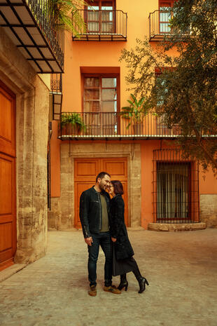 A moment together among warm façades and leafy balconies. A couple leans close together in a courtyard framed by orange walls, wooden doors and wrought-iron balconies.
